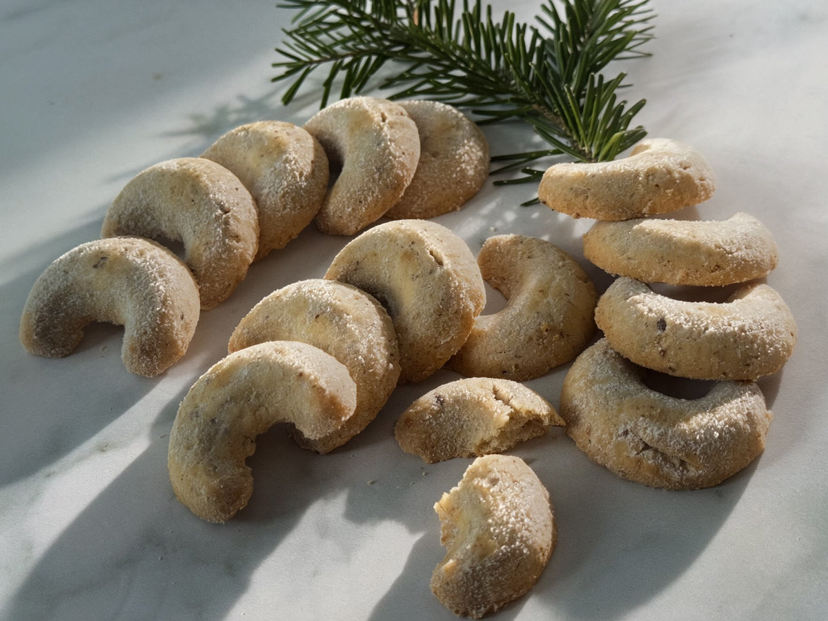 A group of crescent-shaped sugar cookies dusted with powdered sugar, arranged on a light marble surface with a green pine branch in the background.