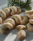 A group of crescent-shaped sugar cookies dusted with powdered sugar, arranged on a light marble surface with a green pine branch in the background.