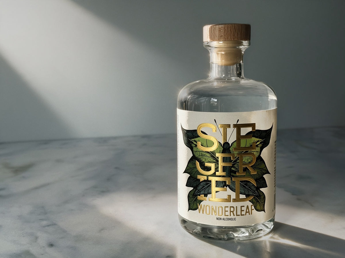 A clear glass bottle of Siegfried Wonderleaf non-alcoholic spirit with a white label featuring gold and green leaf graphics, sealed with a wooden stopper, set on a marble surface with dramatic diagonal sunlight.