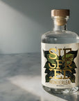 A clear glass bottle of Siegfried Wonderleaf non-alcoholic spirit with a white label featuring gold and green leaf graphics, sealed with a wooden stopper, set on a marble surface with dramatic diagonal sunlight.