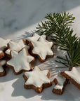 Star-shaped cinnamon and nut cookies with white icing, arranged on a light marble surface next to a green pine branch.