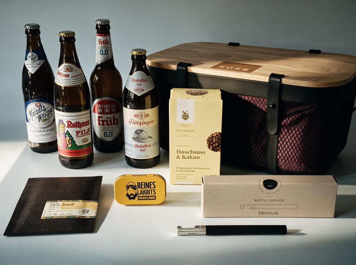 A curated beer gift set arranged on a dark surface with dramatic side lighting. Visible items include four large beer bottles, a wooden-lidded storage case, a black blomus bottle opener with gift box, a slab of chocolate in a black envelope, a small yellow Liquorice tin, and a yellow cookie box.