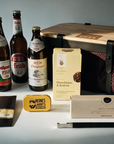 A curated beer gift set arranged on a dark surface with dramatic side lighting. Visible items include four large beer bottles, a wooden-lidded storage case, a black blomus bottle opener with gift box, a slab of chocolate in a black envelope, a small yellow Liquorice tin, and a yellow cookie box.