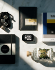 A styled flat-lay of multiple products on a white marble surface with dramatic diagonal shadows. The items include a black drawstring pouch with silver ice cubes, a slab of chocolate in a black envelope, a Bauhaus book, a black notebook, a small black Liquorice tin, and a clear glass bottle with a cork and green botanical label.