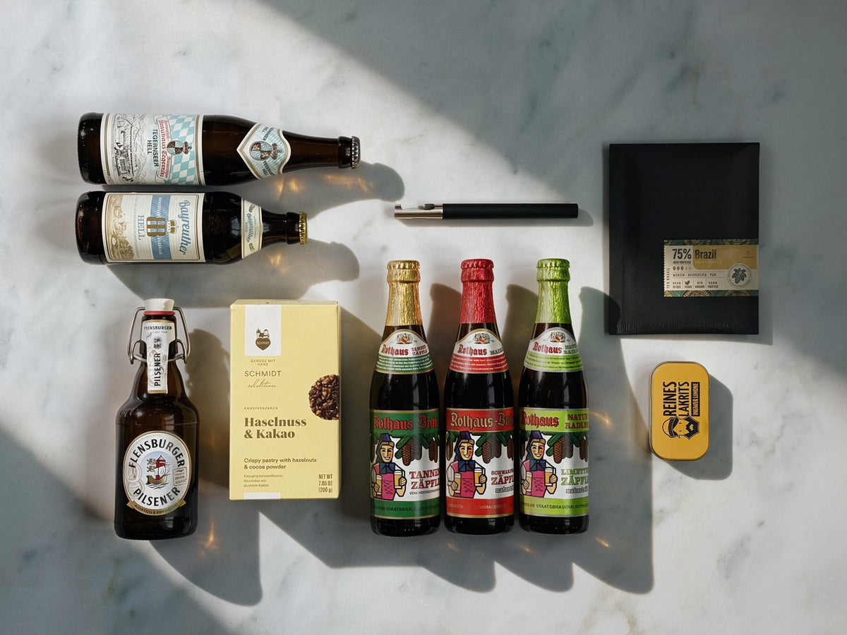 A styled flat-lay of beer bottles and accessories arranged on a white marble surface with dramatic diagonal shadows. Visible items include two dark brown bottles with labels, a clear bottle with a white label, three smaller bottles with colourful labels, a black blomus bottle opener, a slab of chocolate in a black envelope, a small yellow Liquorice tin, and a yellow cookie box.