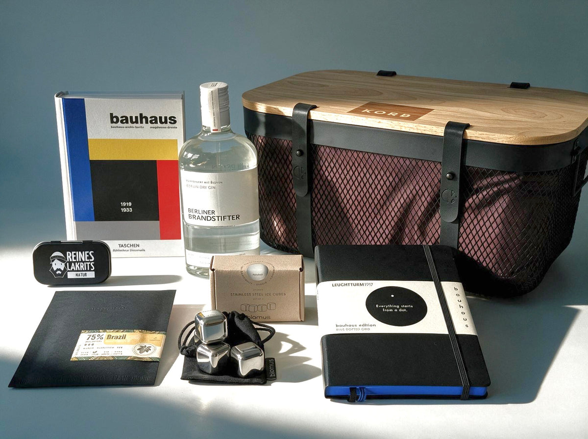 A curated collection of Bauhaus-inspired products arranged on a light surface against a background, including a black mesh basket with wooden lid and liner bag, a Bauhaus book, a gin bottle, a slab of chocolate in a black envelope, liquorice tin, a notebook, and stainless steel ice cubes.