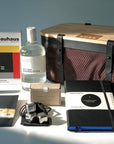 A curated collection of Bauhaus-inspired products arranged on a light surface against a background, including a black mesh basket with wooden lid and liner bag, a Bauhaus book, a gin bottle, a slab of chocolate in a black envelope, liquorice tin, a notebook, and stainless steel ice cubes.