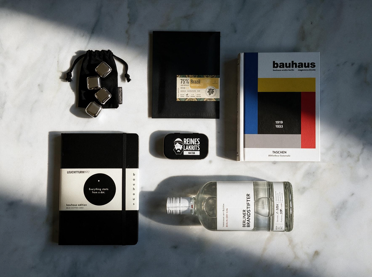 A styled flat-lay of multiple products on a white marble surface with dramatic diagonal shadows. The items include a black and white Leuchtturm1917 notebook, a clear glass bottle of Berliner Brandstifter gin, a Bauhaus book, a small black liquorice tin, a black pouch with stainless steel ice cubes, and a slab of chocolate in a black envelope.
