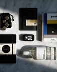 A styled flat-lay of multiple products on a white marble surface with dramatic diagonal shadows. The items include a black and white Leuchtturm1917 notebook, a clear glass bottle of Berliner Brandstifter gin, a Bauhaus book, a small black liquorice tin, a black pouch with stainless steel ice cubes, and a slab of chocolate in a black envelope.