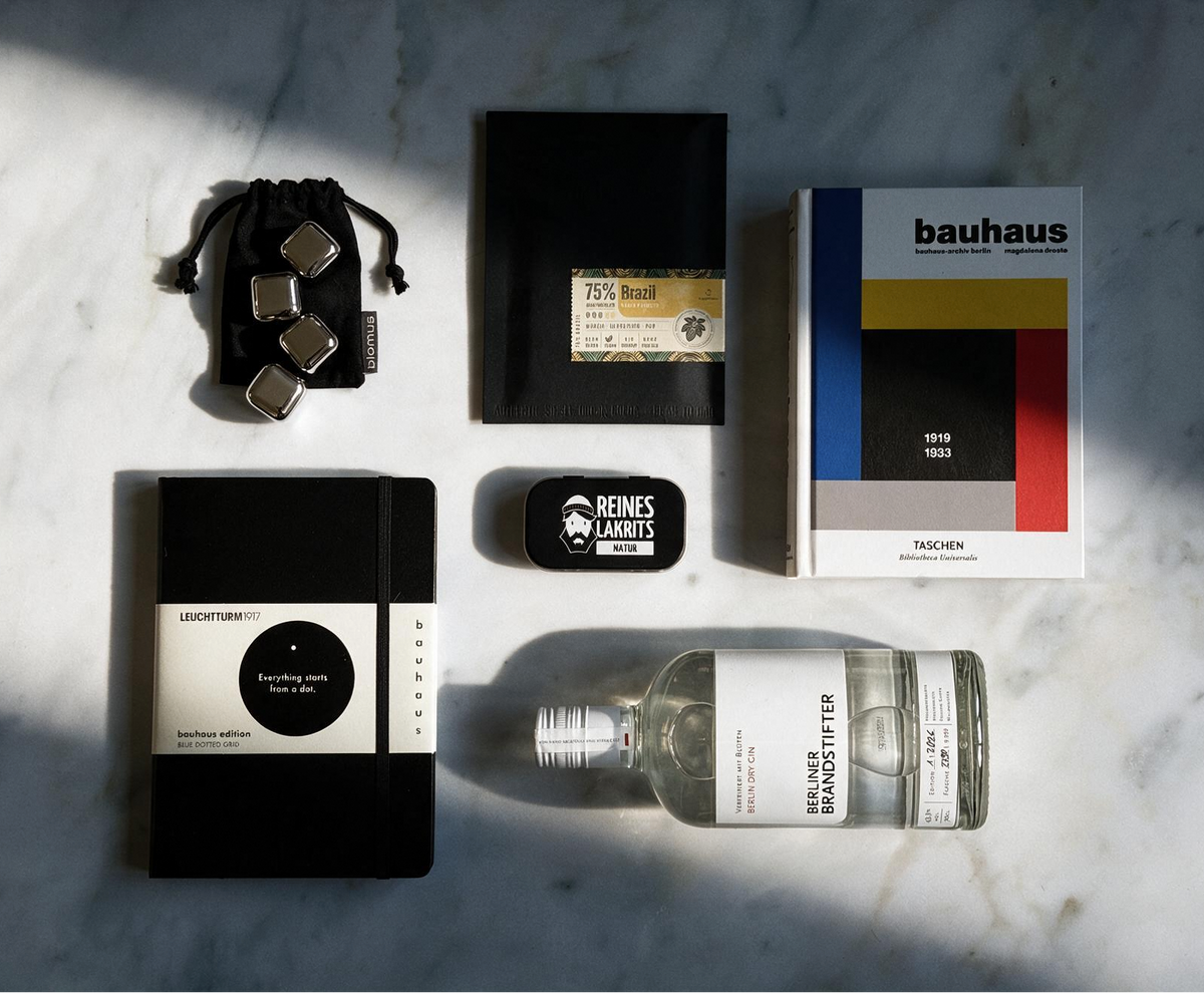 A styled flat-lay of multiple products on a white marble surface with dramatic diagonal shadows. The items include a black and white Leuchtturm1917 notebook, a clear glass bottle of gin, a Bauhaus book, a small black liquorice box, a black pouch with silver ice cubes, and a small black-packaged chocolate.
